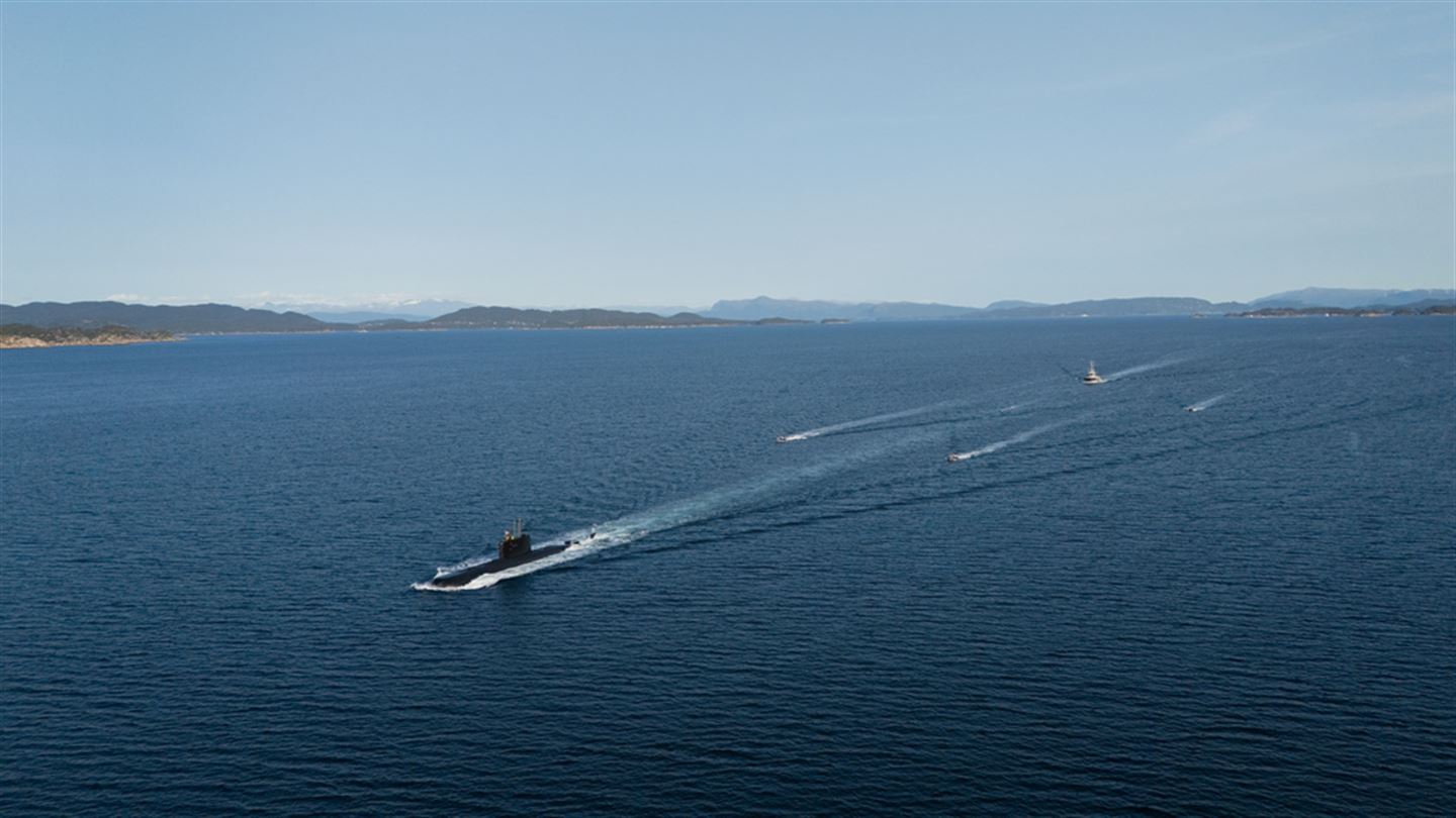 Ubåt seglar foran fleire fartøy i eit norsk fjordlandskap i solskin, sett ovanfrå. 