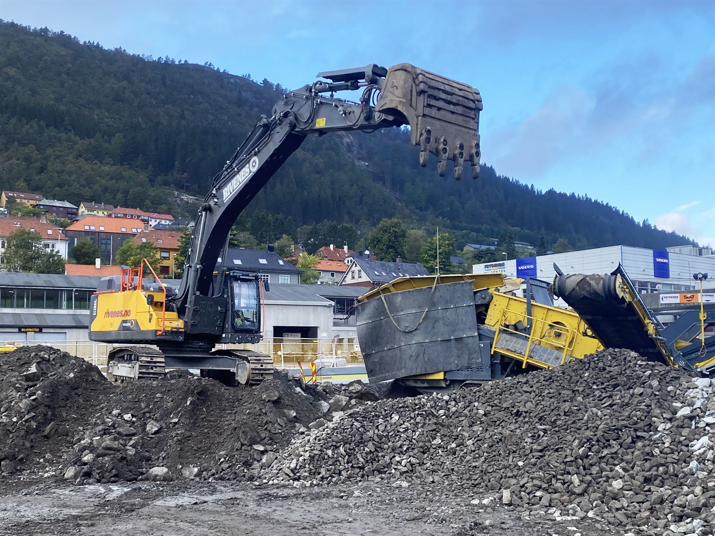 Ein gul og svart gravemaskin står oppå ein haug med stein, og har grabben i luften. Ved sidan av står ein gul og svart maskin som knuser asfalt, også denne på toppen av ein liten steinhaug. 