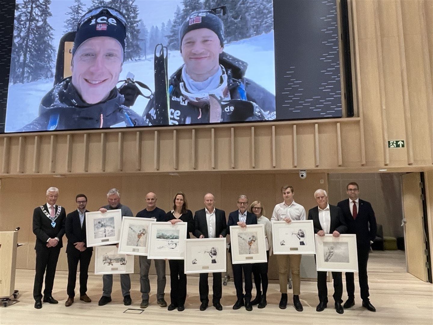 foto av mange folk på rad, dei held eit maleri kvar i handa. på skjerm bak dem er to personar.