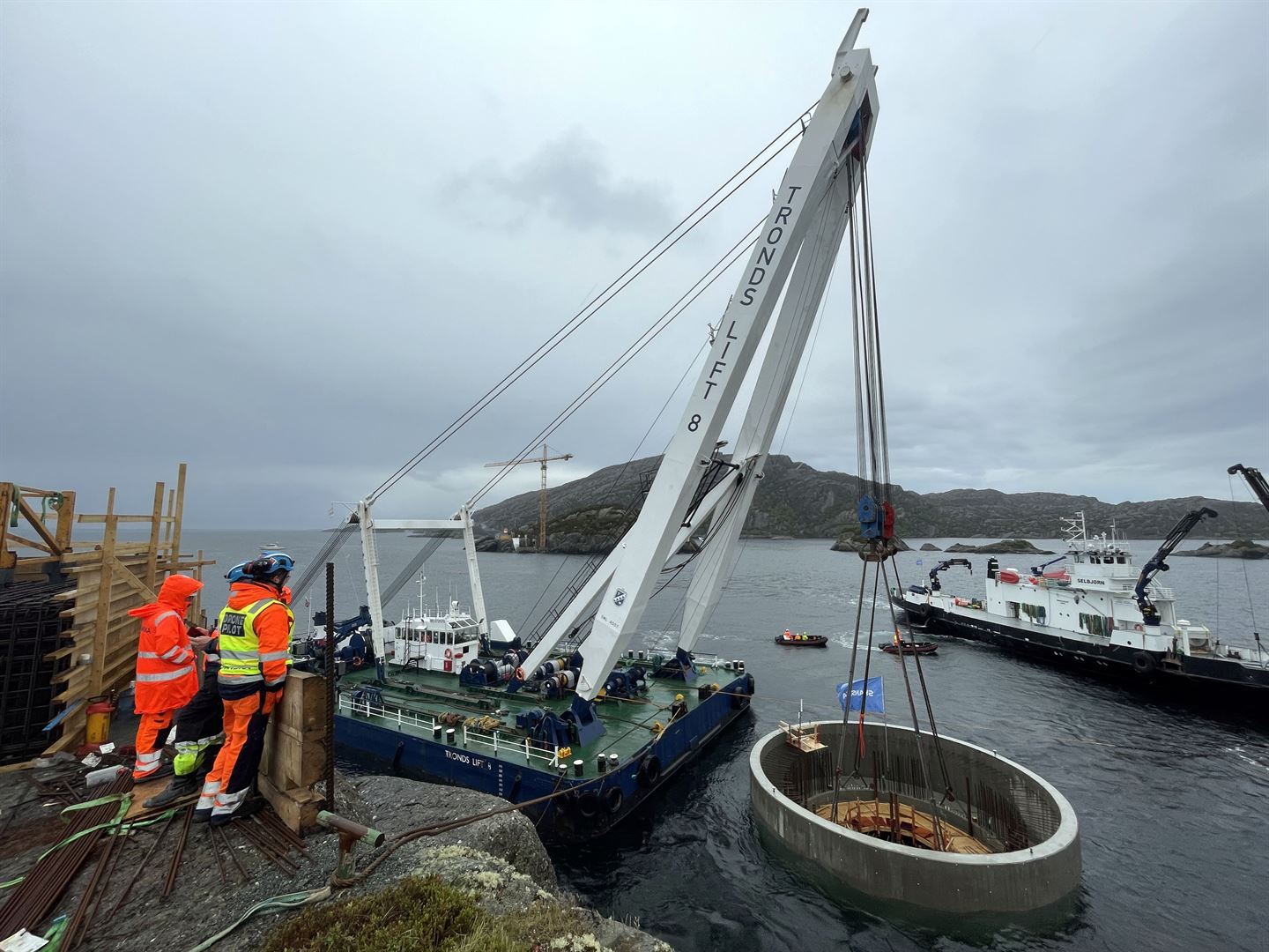 Frå ei holme på kysten ser vi ut mot ein holme litt lenger ut i havet. To arbeidarar i oransje verneklede står i forkant på næraste holmen, dei ser på eit kranskip som set på plass eit stort, rundt betongfundament til ei søyle til ei bru som skal byggast over til den andre holmen. Ei svart og kvit lasteferje og eit par småbåtar med fleire vernekledde arbeidarar hjelp til rundt brufundamentet i sjøen. Gråvèr med grå himmel og grått hav. 