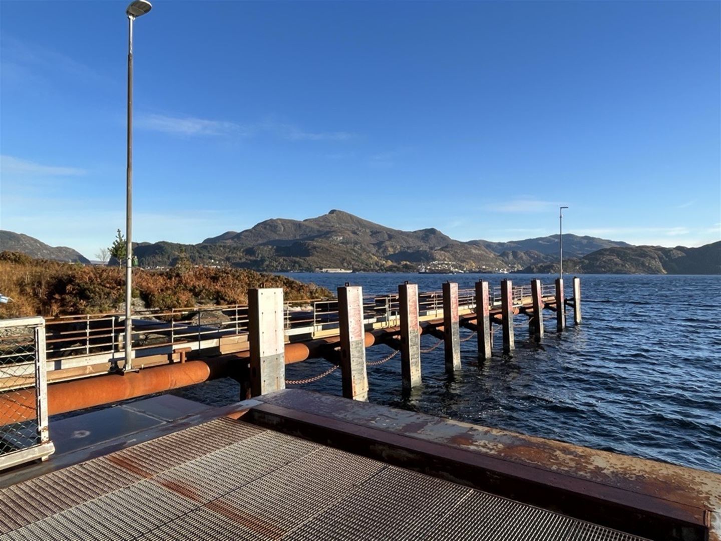 Ferjekai i forgrunn av bildet, med mykje rust. Blå sjø og blå himmel i bakgrunn, holmar og skjer i sjøen.