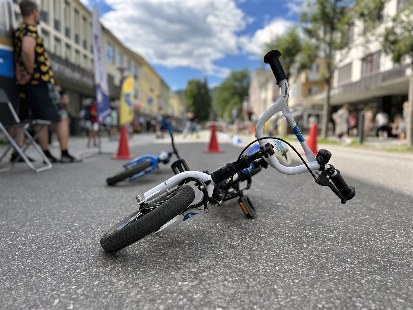 Ein barnesykkel ligg på asfalten med styret og forhjulet framst. Sykkelen er kvit og svart. Bak står tre raude kjegler. 