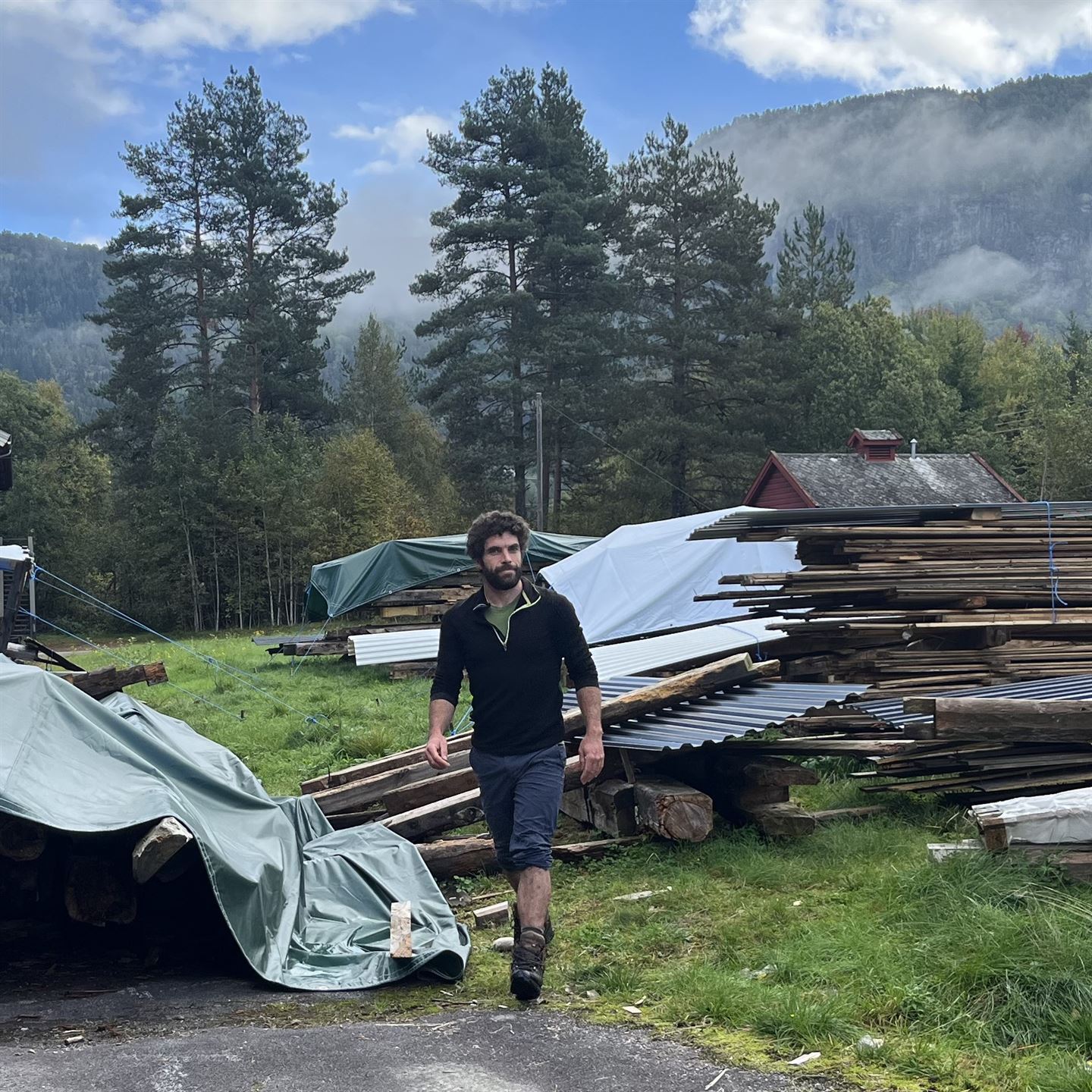 Ein mann med mørkt, krøllete hår, kledd i fritidskle og shorts. Bak han ligg det plank og blikktak stabla oppå kvarandre. Til venstre for han ligg ein grøn presenning. 