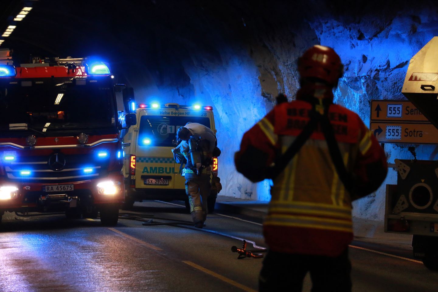 Bildet viser ein brannbil og ein ambulanse med blålys i ein tunell. Ein brannmann bærer ein pasient over skuldra mot ambulansen. Foran i bildet står ein brannmann med ryggen til. 