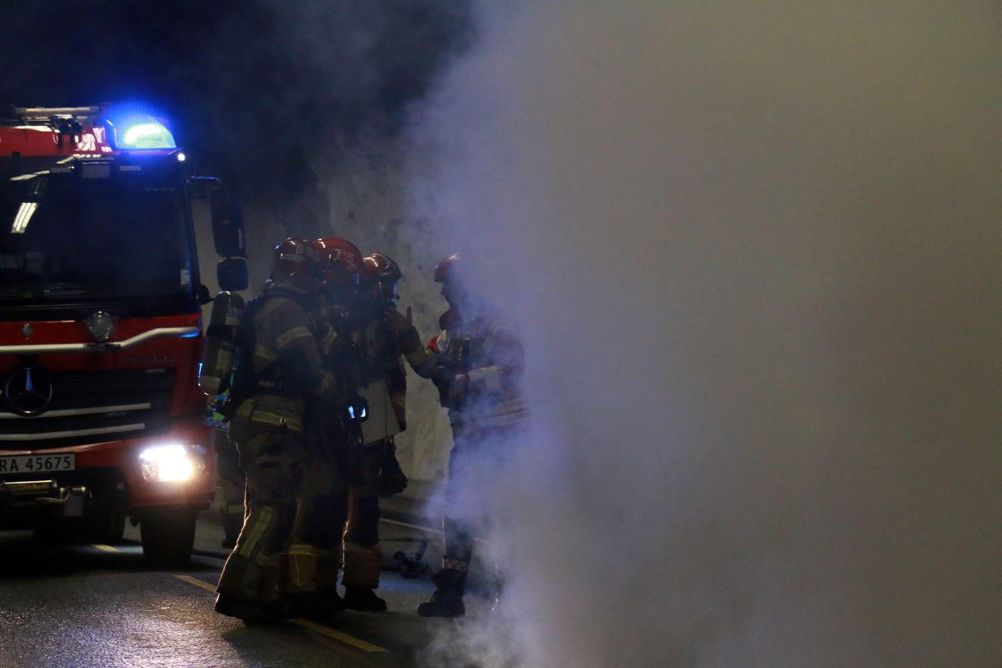 Bildet viser tjukk røyk som delvis dekker fire brannmenn som står i ein tunell. Bak dei står ein brannbil med blålys på. 