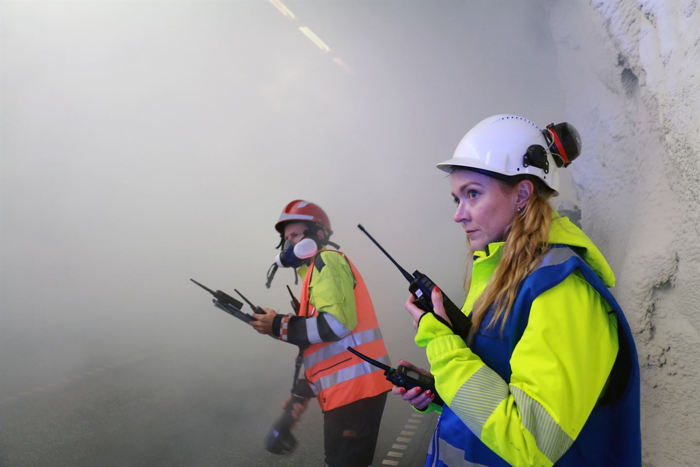 Bildet viser brannvernleiar Marie Svendal Hatlen og ein brannmann. Dei står begge i tjukk røyk inne i ein tunnel. Marie har kvit hjelm og gul jakke på. Ho har to walkietalkier, ein i kvar hand. Brannmannen bak ho har raud hjelm og maske på. 