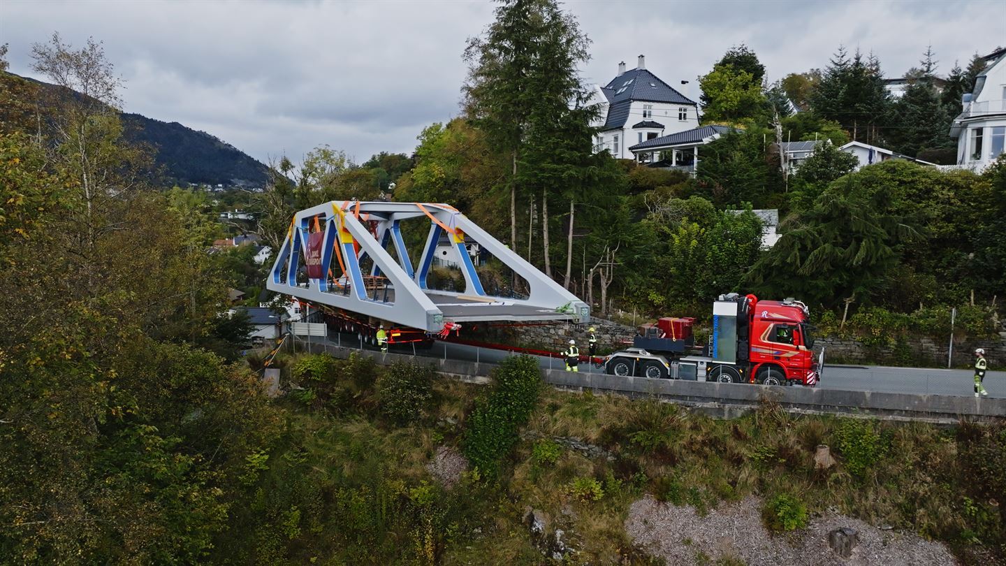 Bilder viser en gangbro fra siden. Broen er blå og grå og blir kjørt på et kjøretøy med et rødt  førarhus. Brua svinger seg utfor vegen. 