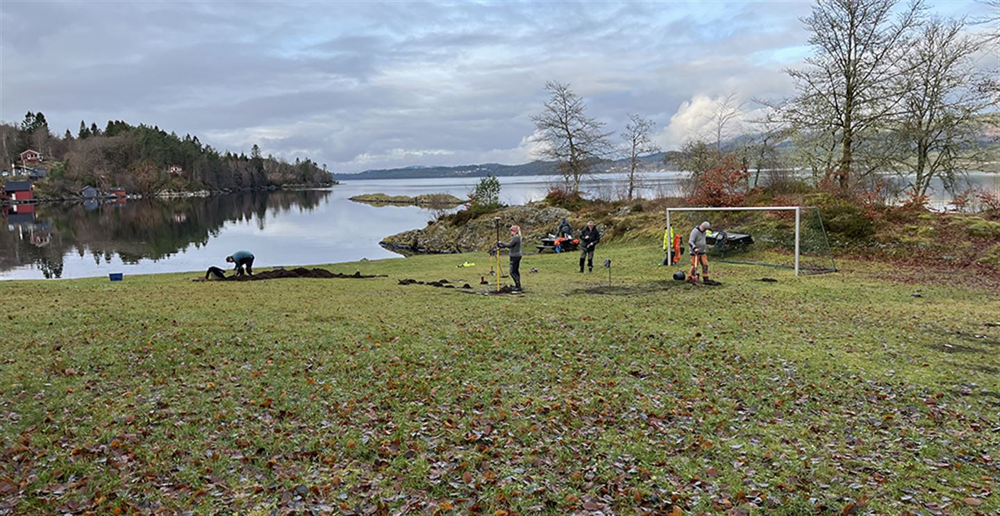 Arkeologar i arbeid på flat grasmark med eit fotballmål, som ligg ved sjøen.