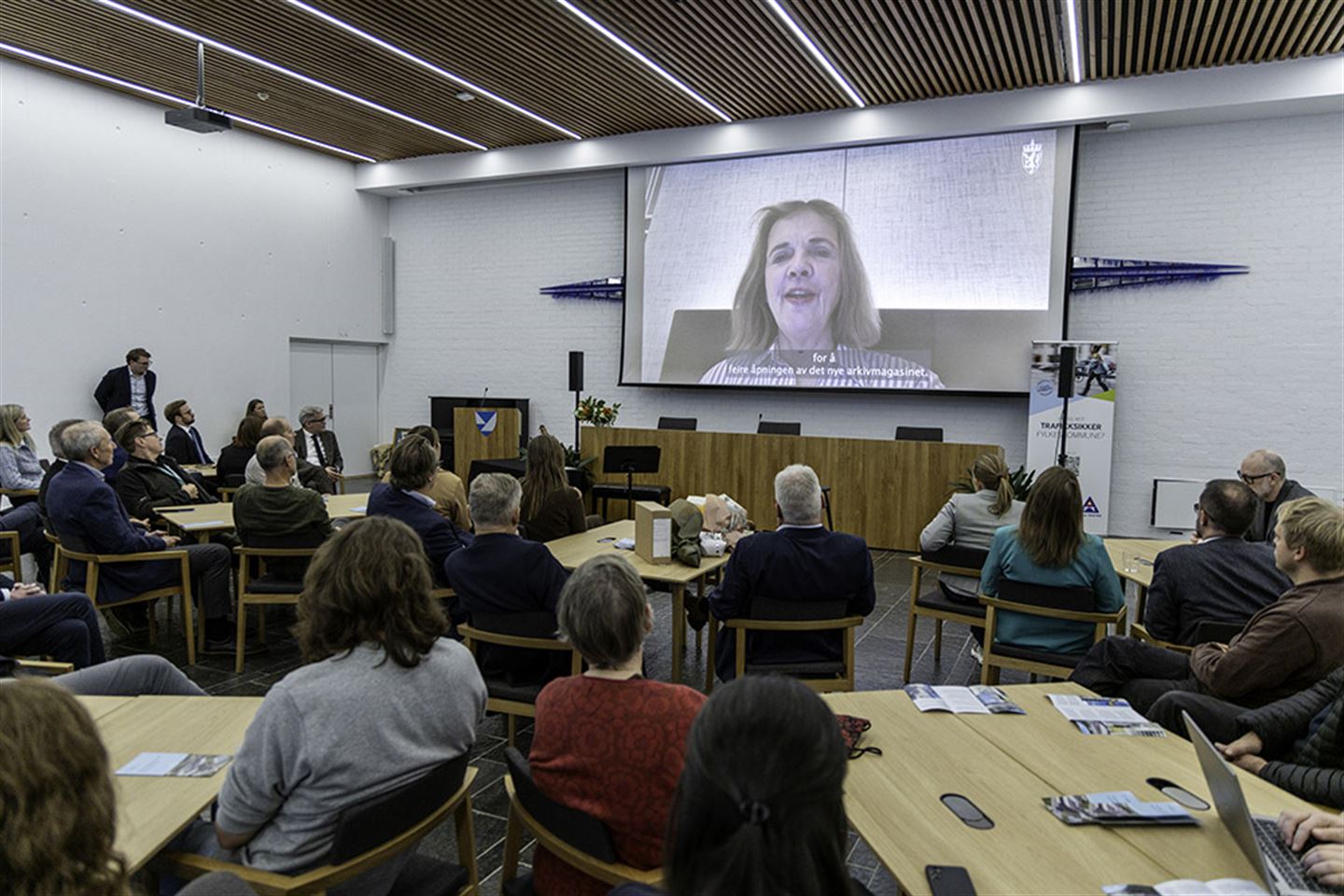 Kvinne på storskjerm framfor forsamling i Firdasalen på fylkeshuset på Leikanger.