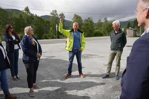 Ein mann med kort, lyst hår, blå genser og gul refleksjakke held armen i været. Ved sida av står ein mann med grått hår og grøn genser. Rundt dei står det fleire menneske og høyrer på. Foto: Silje Alvsaker