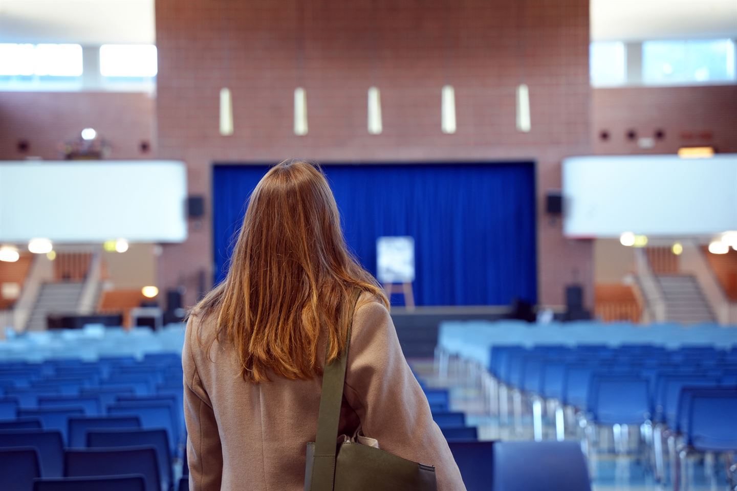 Ei dame med brunt hår, grå kåpe og veske over skuldra. Ho står med ryggen til fotografen og ser utover ein sal med stoler, ei scene med blå gardiner trekt for. 
