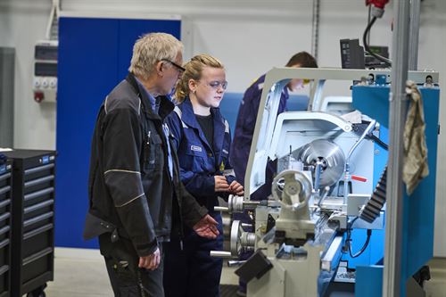 Bilde frå klasserom på yrkesfag på Førde vidaregåande skule. Ein lærar kledd i svarte arbeidsklede med kvitt hår og vernebriller står i forkant av bildet og instruerer ein kvinneleg elev i blått arbeidstøy og vernebriller og lyst hår i flette ved ein metall-dreiebenk. Dei to har lys i ansiktet og vi ser metallet gå rundt i dreiebenken. Ein mannleg elev står ved ein dreiebenk i bakgrunnen. Dreiebenken er blå og eit skap i bakgrunnen er blått. Jenta ser konsentrert på metallet som blir dreia. 