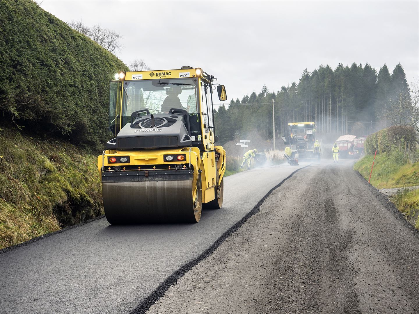 Gult køyretøy jamner ut nylagt svart asfalt. På vegen ved sida av er det grus. Fleire maskinar og menneske er på arbeid eit stykke unna. De går alle i gule kle. 