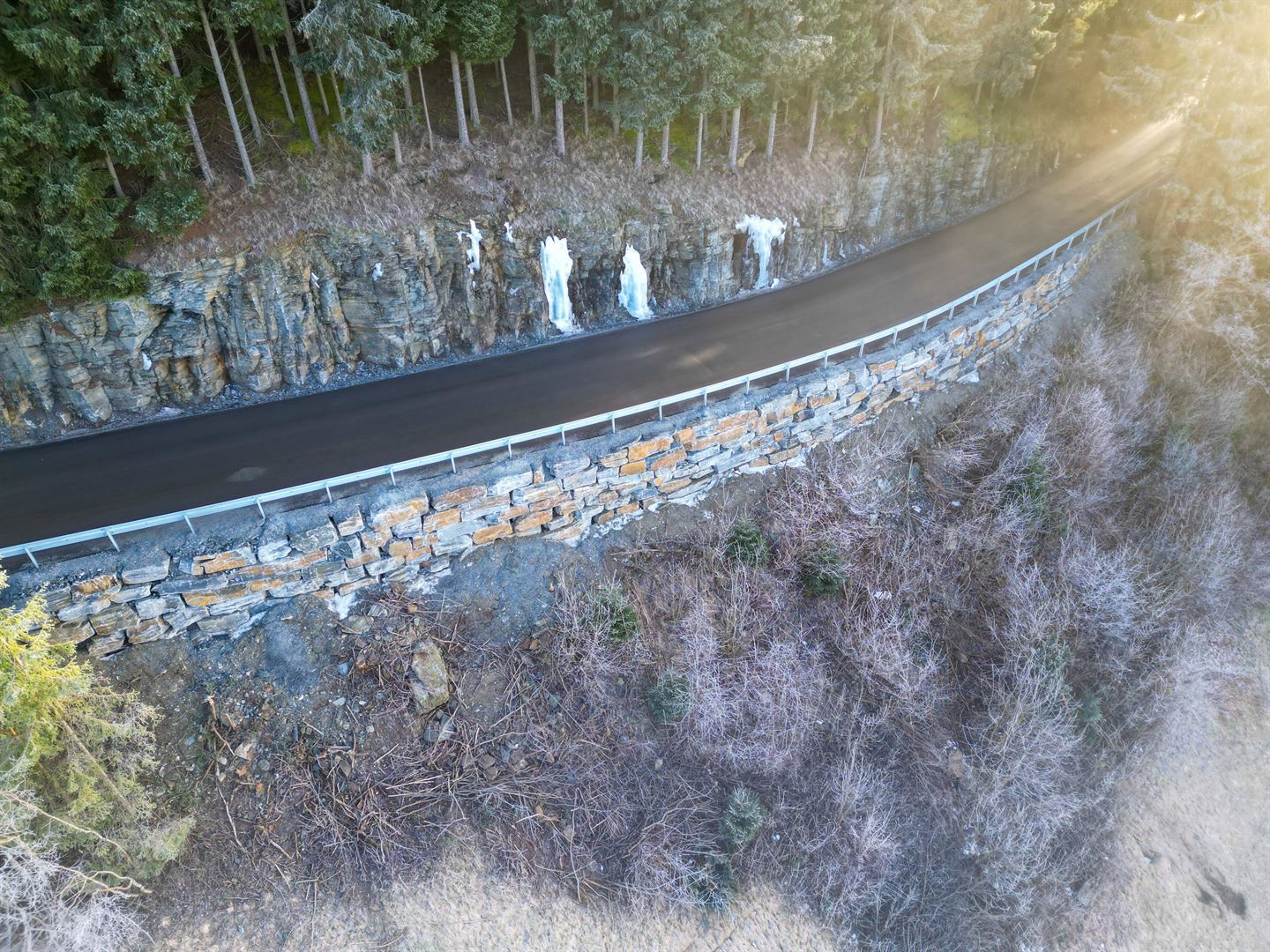 Ein smal, nyasfaltert smal veg med ei køyrebane slyngar seg over ei bergskrent med ny mur i naturstein under vegen nedover stupet og ei skæring i fjellet med granskog over på oppsida av vegen. Det er vinter og issvullar og istappar i skjæringa over vegen. 