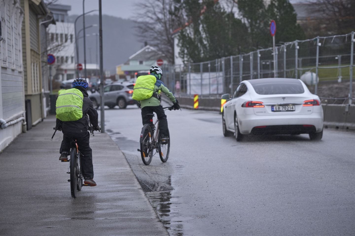 To skuleborn på sykkel langs ein veg med grå asfalt. Begge har på hjelm og gult, reflekstrekk på sekkene sine. Det køyrer ein kvit bil forbi dei to syklistane. Været er grått og det er fleire dammar i vegen. 