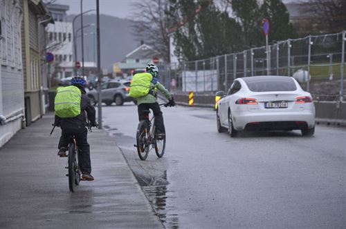 To skuleborn på sykkel langs ein veg med grå asfalt. Begge har på hjelm og gult, reflekstrekk på sekkene sine. Det køyrer ein kvit bil forbi dei to syklistane. Været er grått og det er fleire dammar i vegen. 