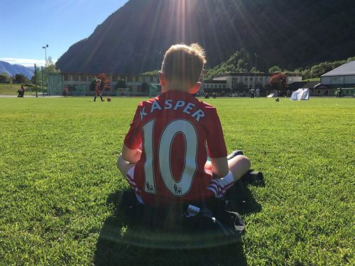 Gut som sit på fotballbane med ryggen til kameraet. På ryggen står det talet 10 og Kasper. I bakgrunnen er det ungar som spelar fotball.