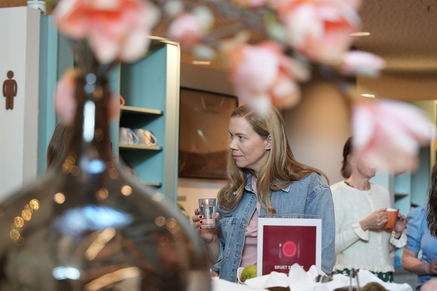 Dame som holder et glass med vann snakker med noen som er skult bak en dekorativ vase.
