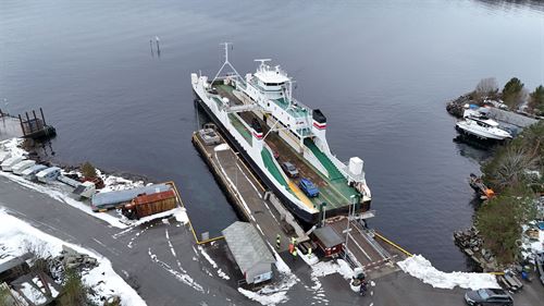 Vi ser ei ferjekai frå lufta med land i forgrunn og grå sjø bak. Ein smal veg går ned til kaia, der ligg det ei ferje inn til ei smal utstikkande kai. Ferjebrua er og smal og lita, og mykje mindre enn det ferja eigentleg treng i høve si breidde. Ferja er lita, kvit med grønt dekke, med sidestilt styrehus. Bommen er nede og 5 bilar er synlege på ferja. det er snø på bakken og mykje rust på kai og ferje. 