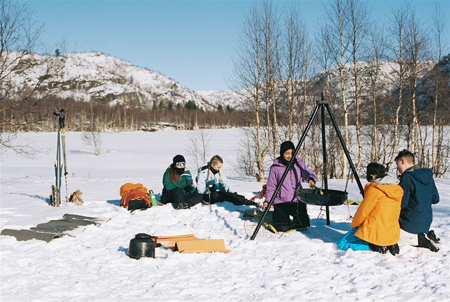 Foto av barn/ungdomar samla rundt bålpanne ute i snøen. 