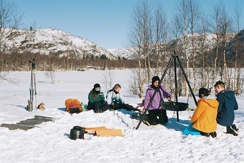 Foto av barn/ungdomar samla rundt bålpanne ute i snøen. 