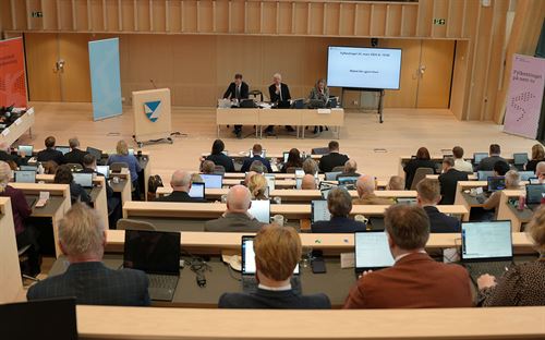 Oversiktsbilde av eit fylkestingsmøte i Vestlandssalen i Vestlandshuset i bergen. Vi ser framover i salen frå bak sitteplassane som går nedover til golvet framme i salen, der er det eit smalt bord med tre personar, og ein talarstol til venstre i bildet. Materialane i salen er i tre og er trefarga. fremst i bildet ser vi ryggen og bakhoda til fire personar som ser framover i salen. 