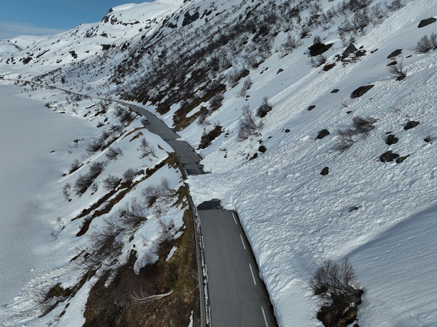 Snøkledd landskap med fjell og eit is og snødekt vatn i forgrunnen. Vegen som går langs vatnet er grå og fri for snø, men eit snøskred har gått ned på vegen frå ei snøkledd fjellside. 
