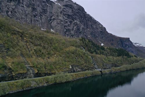 Vi ser ei bratt fjellside som endar i fjorden. Lengst nede ved fjorden går det ein smal veg. Øvste del av fjellsida er berre blankt fjell og stein, noko skog lenger nede over vegen. Grå fjord og overskya kvit himmel. Grønn skog og mørkegrått fjell. 