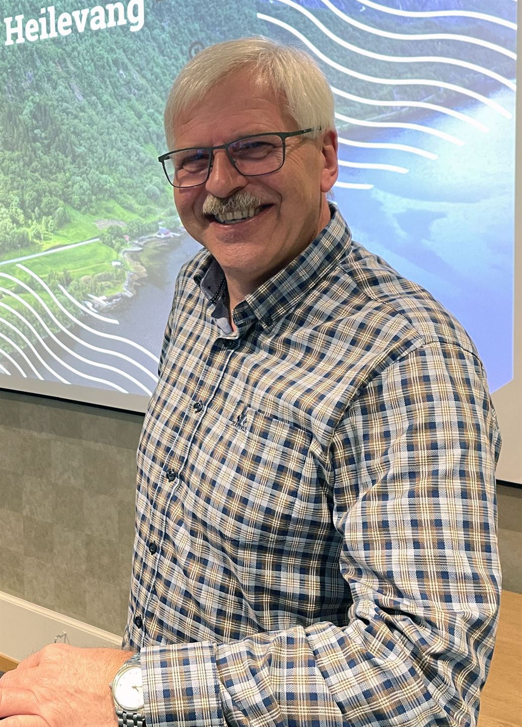 Vi ser ein eldre mann frå livet og opp, han smiler til kamera. Han har grått hår og gråkvit bart, og har briller, rutete skjorte. Bakgrunnen er ein kvit vegg. 