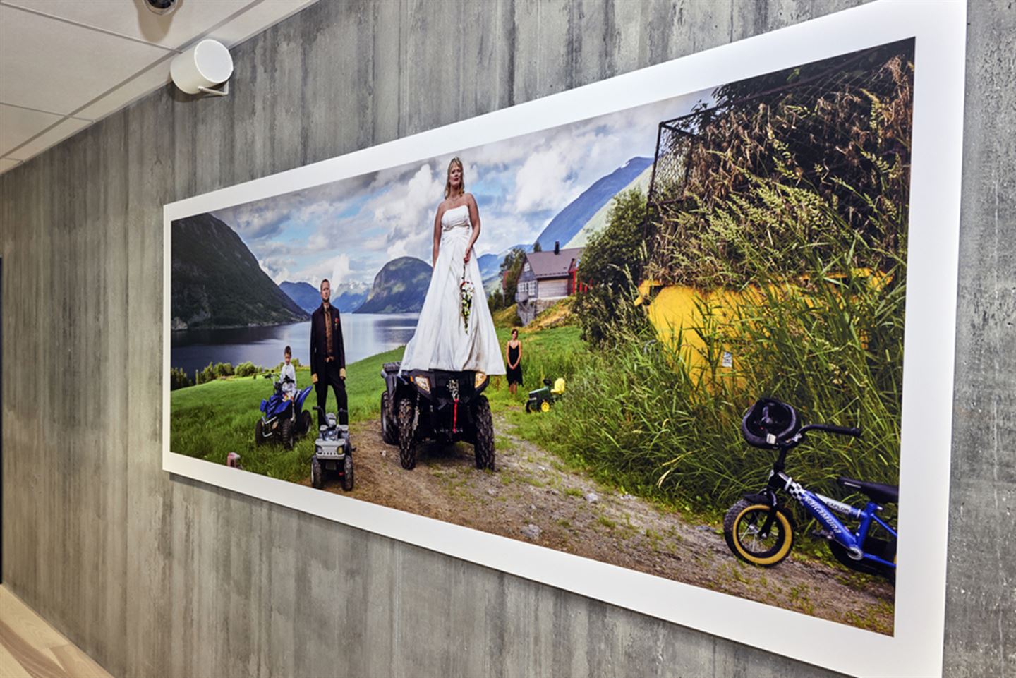 Stort fotografi på betongvegg. Bildet syner ei brur, som står på ein atv, ein brudgom som står ved sidan, og ein gut på ein mindre atv i bakgrunnen.