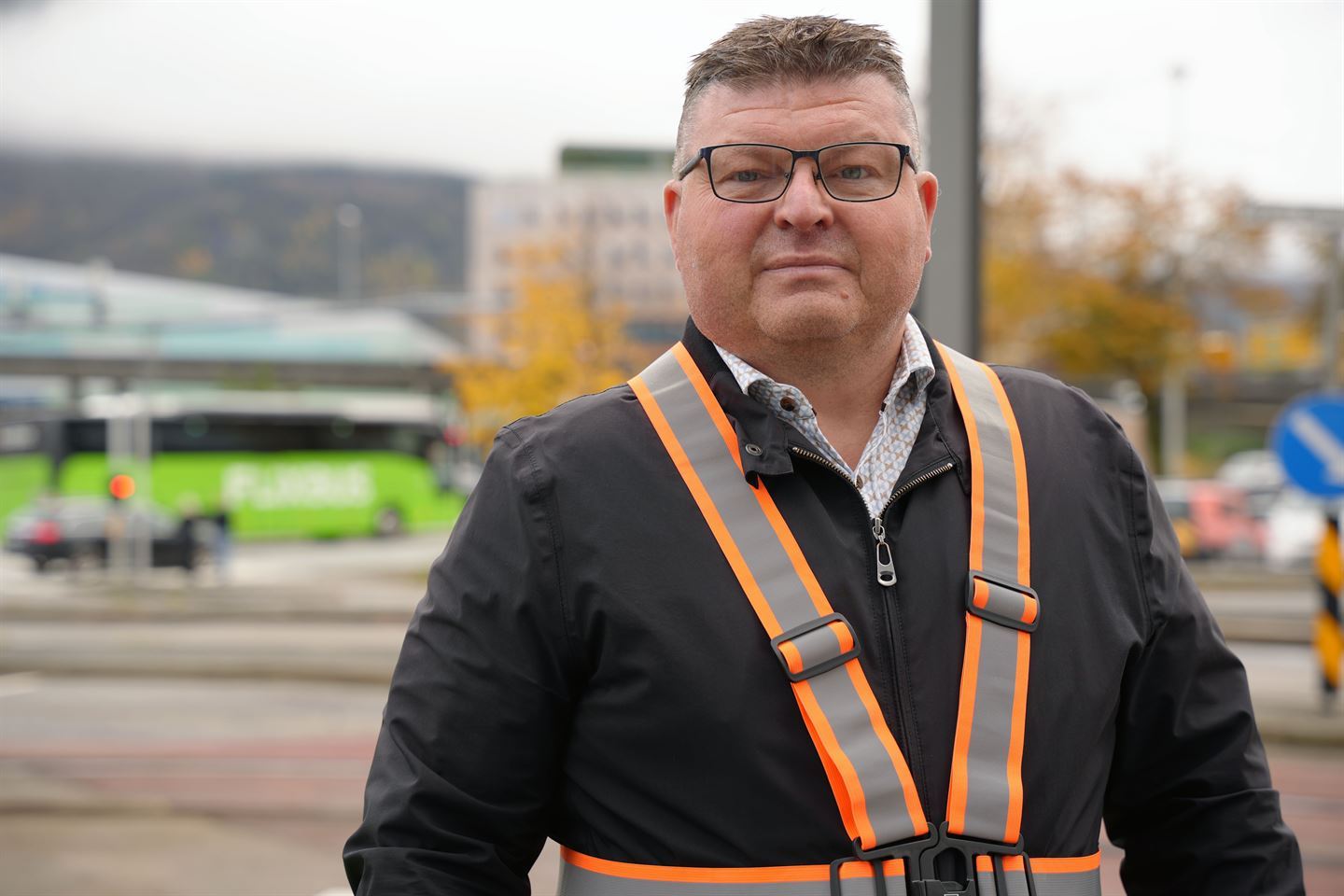 Ein mann med kort, mørkt hår, briller, mørk jakke og oransje refleksband på seg står ute. Bak han er det veg og trafikk. Foto: Silje Alvsaker