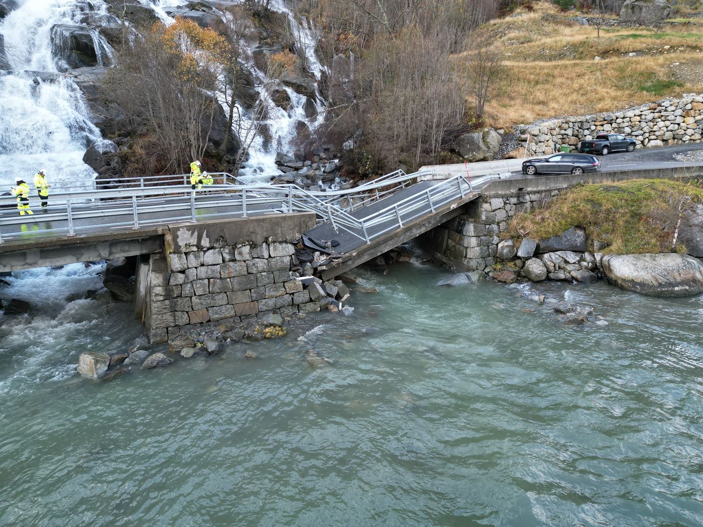Ein foss med kvitt vatn strøymer nedover ei fjellside og under ei øydelagt bru. Vatnet i fjorden er grønt. 