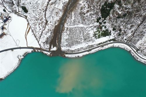 Ei bratt fjellside dekt i snø. I midten av fjellsida har eit ras gått som har tatt med seg vegen nede ved fjorden. Vegen går langs ei fjord med grønt vatn. 