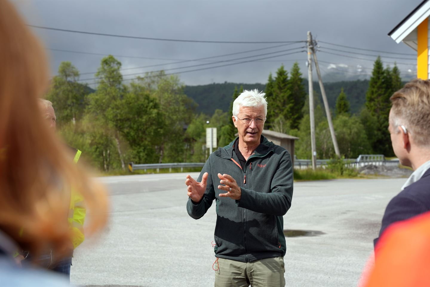 Ein mann med lyst grått hår, briller med mørk fleecejakke og grøn bukse. Bak han er ein stor asfaltert plass og ein veg med autovern. Foto: Silje Alvsaker (Vestland fylkeskommune)