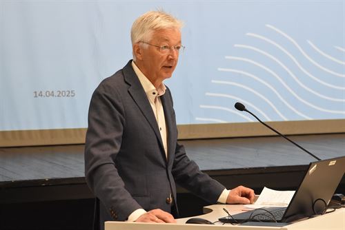 Ein mann med grå dress og lyst grått hår står på ein talarstol og se utover ein forsamling. 