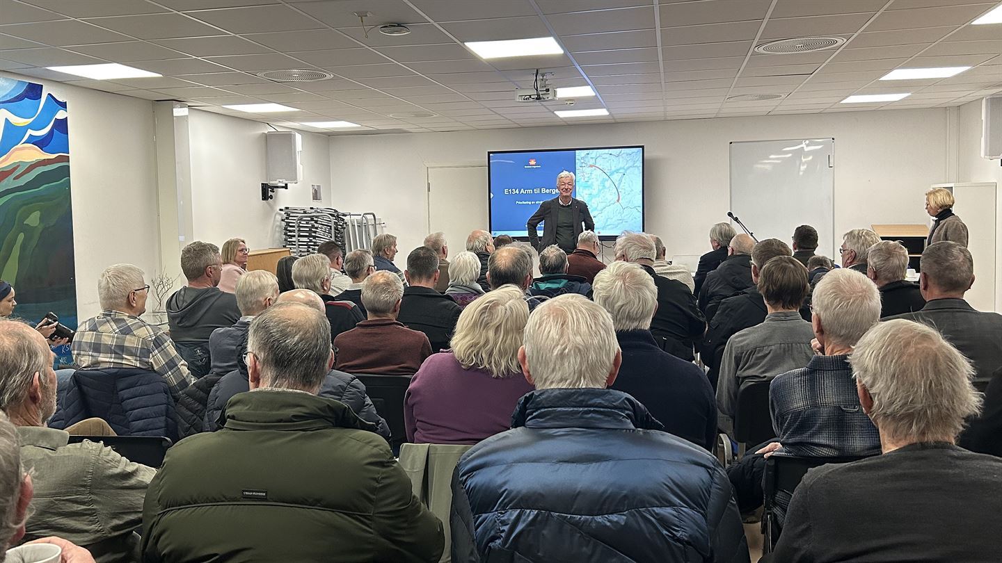 Ein mann med lyst, grått hår står foran ein forsamling av folk som sit og høyrer på. 