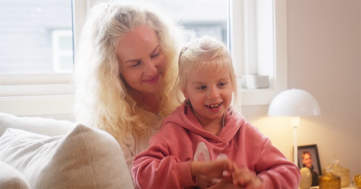 Ei dame med lyst krøllete hår har dattera på fanget. Jenta har lyst, stritt hår, rosa hettegenser og smiler. 