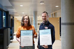 Ei dame med oransje genser, halvlangt hår og briller står ved sida ein mann i mørk fleecejakke. Han har ein buff i halsen, kort hår og skjegg. Dei to held kvar sin ramme med eit diplom der det står "Trafikktryggingsprisen i Vestland 2025". Foto: Silje Alvsaker/Vestland fylkeskommune