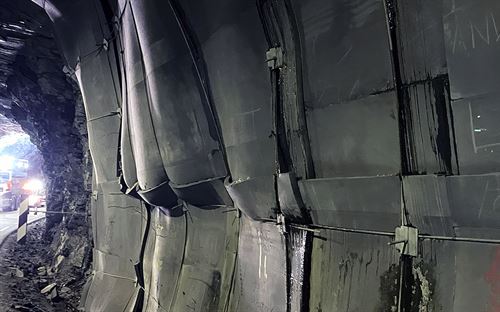 Ein tunellvegg er dekka av dårleg festa grå plater festa med boltar, over ein grå smal dårleg asfaltert veg. Vi ser så vidt utgangen av tunellen og ein bil som står utanfor til venstre i bildet. Over platene er det grovt hakka fjellvegg. Fleire plater bular ut, noko bak dei sprenger seg ut. 