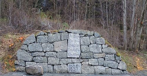 Steinmur med inskripsjon i stein, heller framfor og ei vassblokk ståande på hellene. 