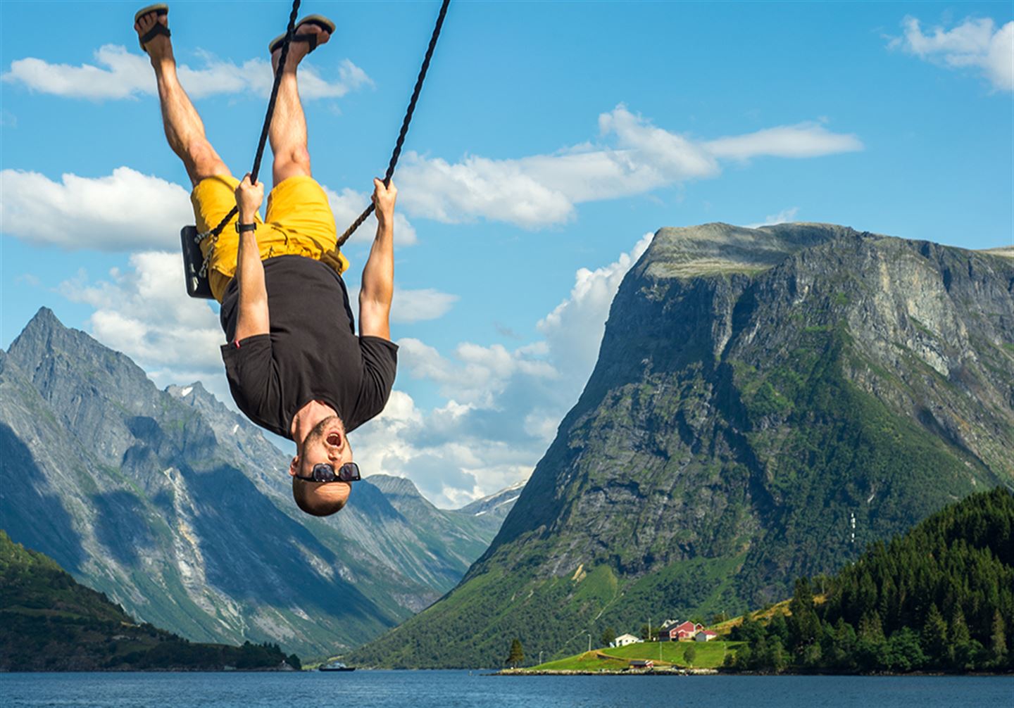 Ein mann som heng opp ned og gyngar på ei huske med ein fjord i bakgrunen