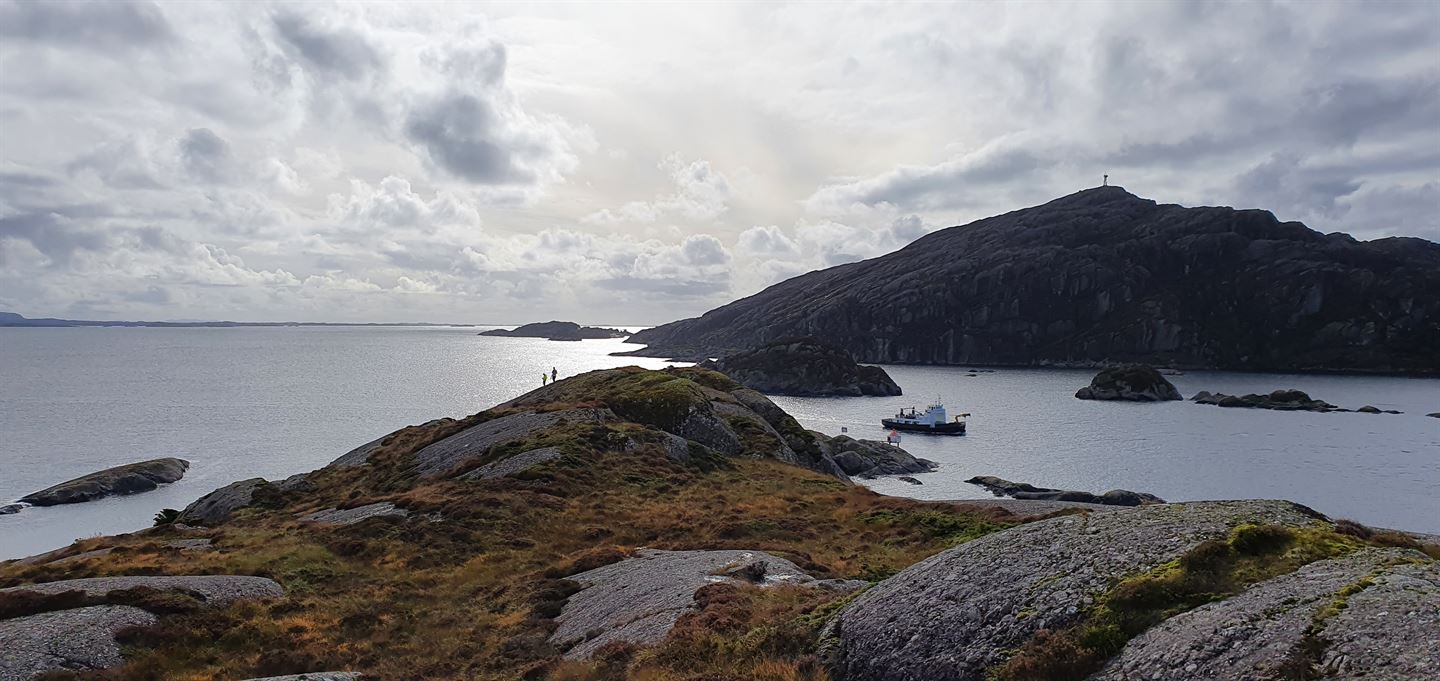 Kystlandskap med holmar og eit sund. Ein fiskebåt passerer gjennom sundet. Midt i bildet ligg Håfjellet, med ei mast på toppen.