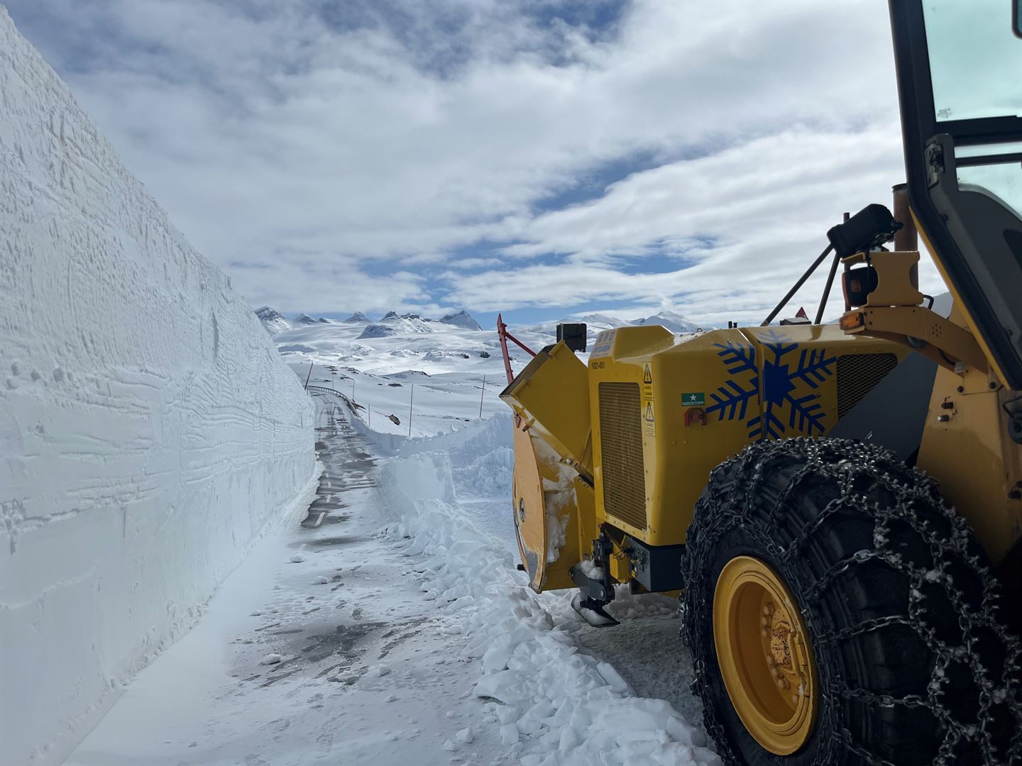 Snødekt høgfjellslandskap, ein veg snor seg fram gjennom bilde med ein oransje brøytebil i forkant av bildet. Bak den delvis snødekte vegen ser vi eit fjellmassiv som stikk opp av snødekt bre. Høg brøytekant på venstre side av vegen fremst i bildet. blå himmel og kvite skyer. 