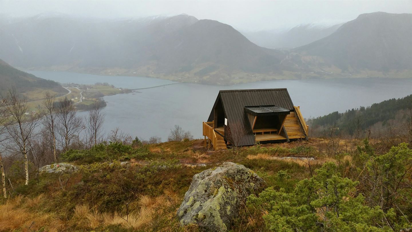 Bilde av ei dagsturhytte. I bakgrunnen er det Jølstravatnent og fjell. Regntung dag. 