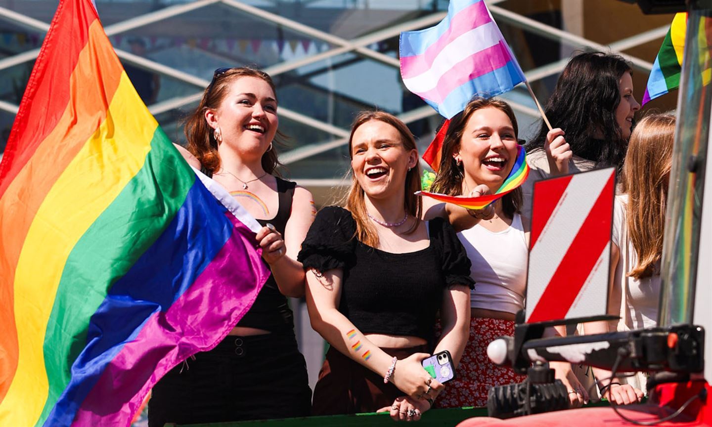 Nærbilde av tre unge kvinner som står i lag. Dei held pride-flagg, smiler og ler, og har pride-flagg måla på armane. Sola skin og dei er sommarkledde. Urban bygning i bakgrunnen. 