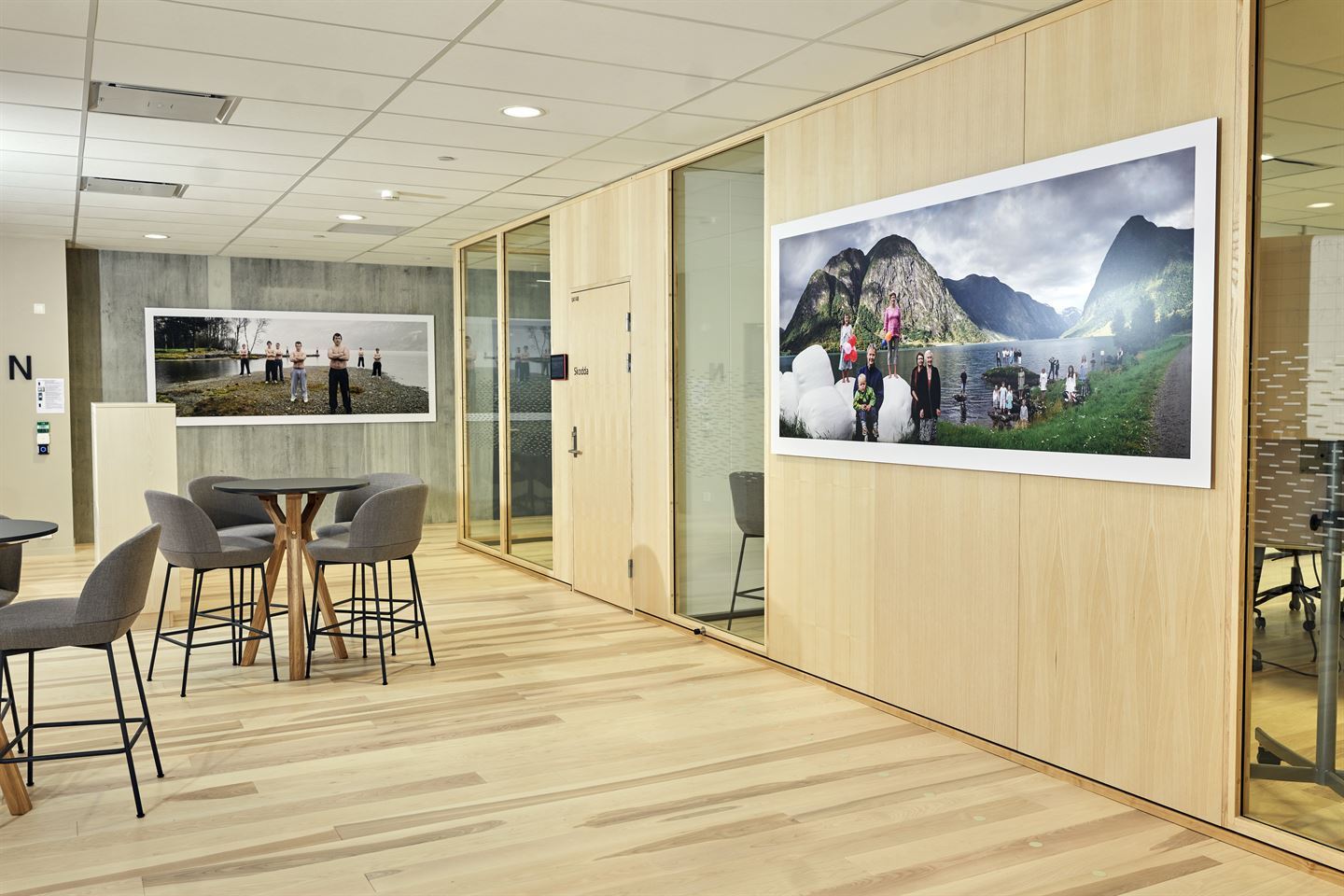 To fotografi på to ulike veggar i kontorlandskap med møterom. Motiva er menn i bar overkropp og menneske og rundballar langs eit vatn.