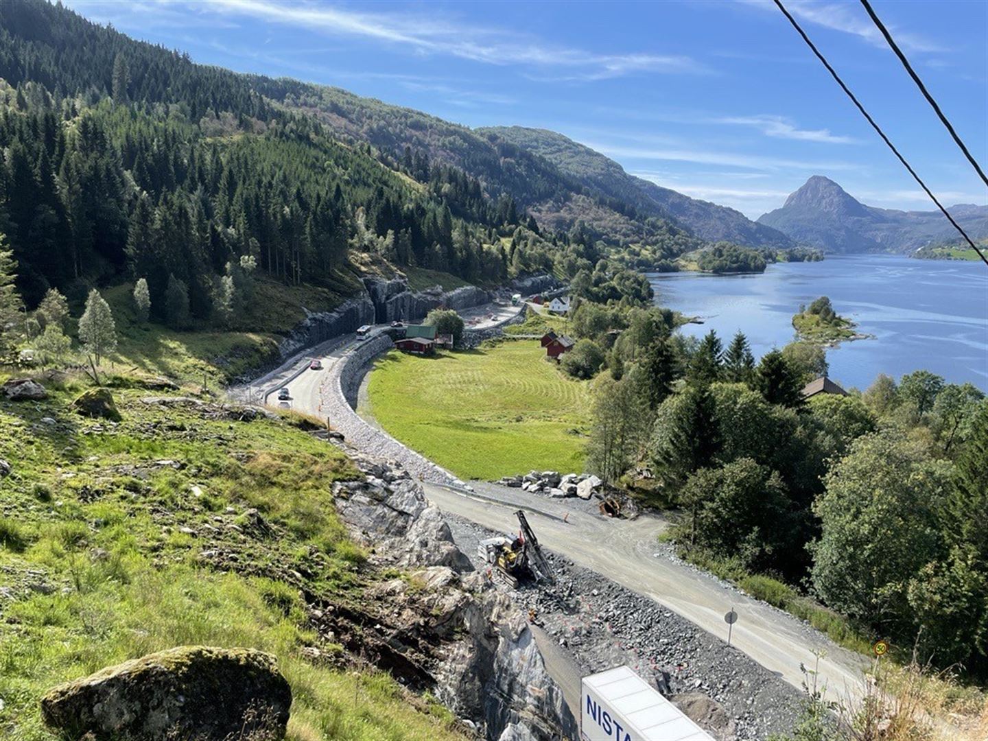 Bilde frå vegbygging. Vegen går langs bratte fjell ved fjord, mykje mur og grøne bøar. Sol og sommar.