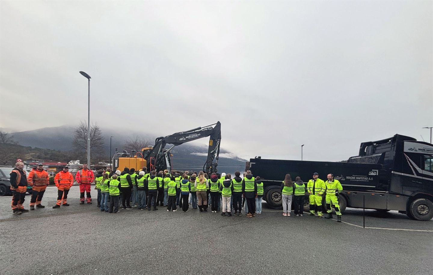 Mange ungdommar står med ryggen til fotografen iført gule refleksvestar, framfor ein lastebil og ei gravemaskin. På sida av dei står personar i gule og oransje vernekle.