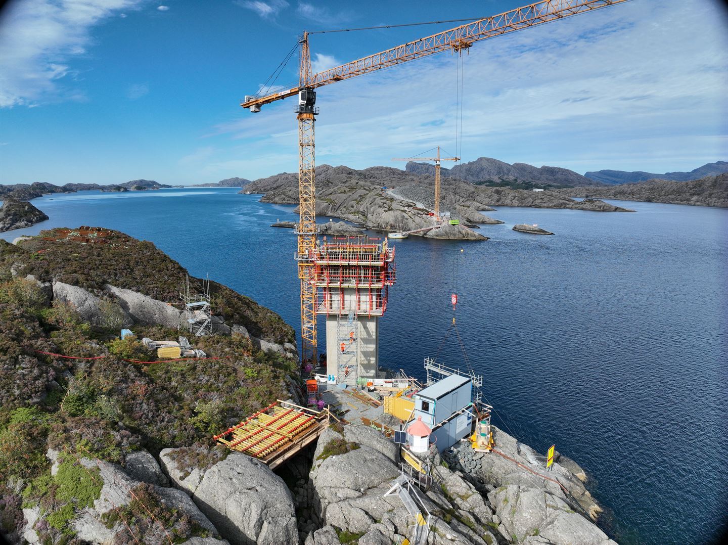 På ein holme i sjøen står det to pilarar i betong, med ei forskalingskasse på toppen. Ved sidan av står ein lift, og på bakken er det diverse utstyr, plattingar, trapper og to grå containerar, den eine på toppen av den andre. 