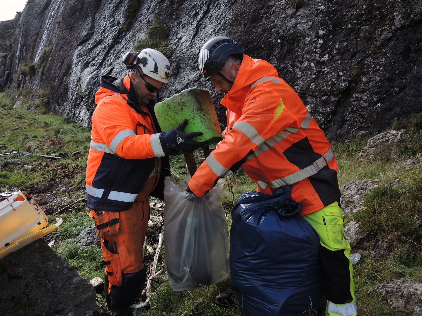 To menn med hjelm i oransje vernekle. Den eine held ein søppelsekk, medan den andre tømmer innhaldet i ei bytte over i sekken.